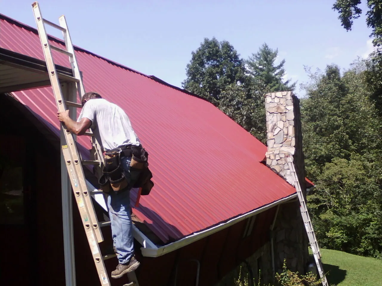 Roofer on a ladder beside a red metal roof and stone chimney in Sutersville